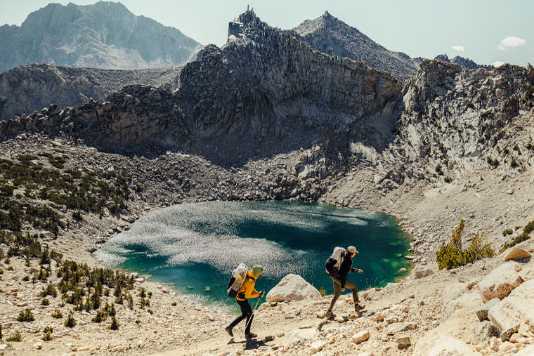 Two hikers with backpacks standing near a lake surrounded by mountains.