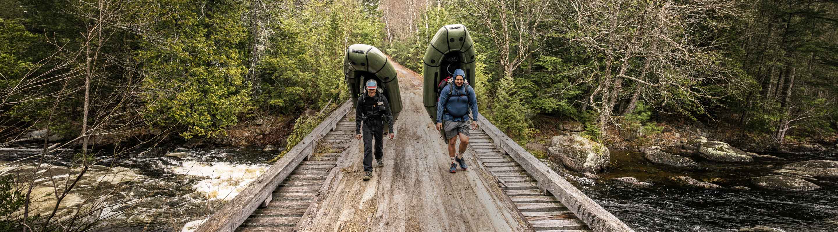 Two hikers carrying rafts crossing a wooden bridge over a river in a forest.