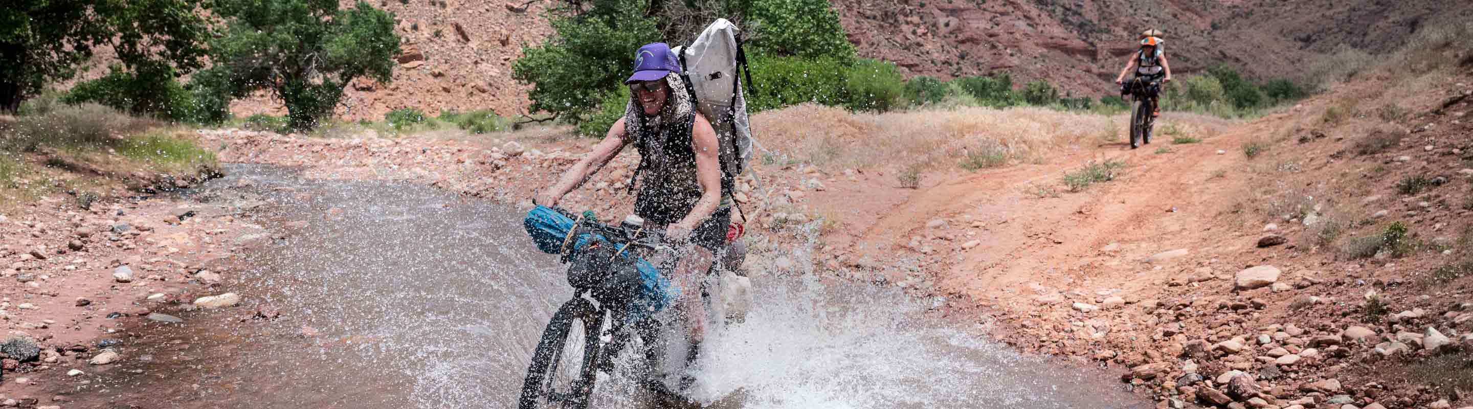 Person riding a bike through water on a dirt road with mountains in the background