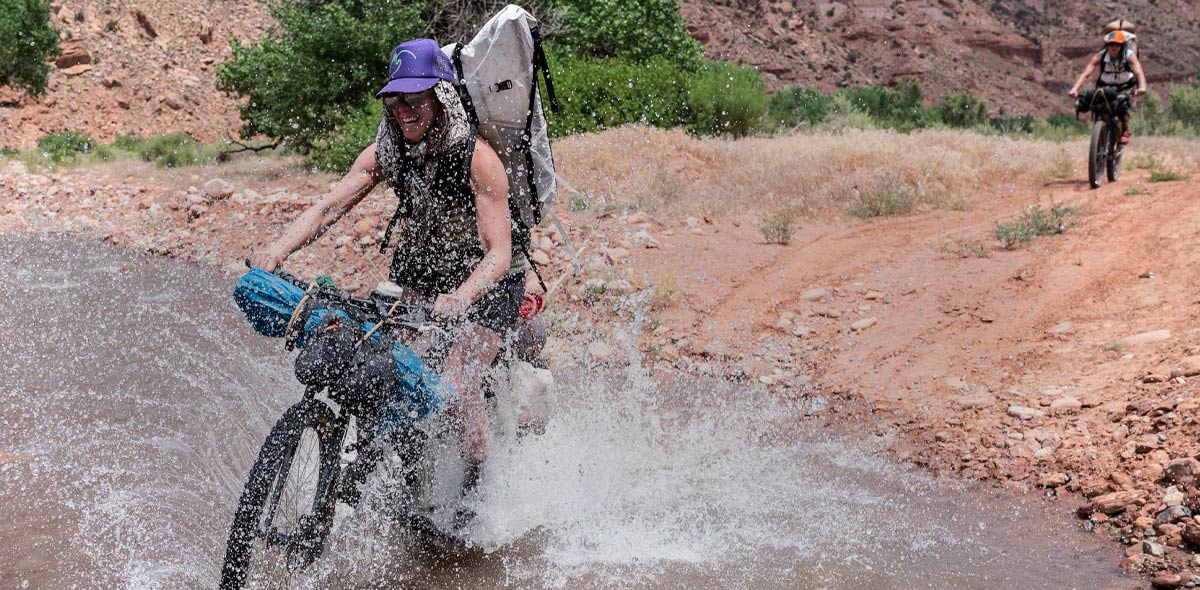 Person riding a bike  through water wearing a camping gear backpack on a dirt road with another person in the background.