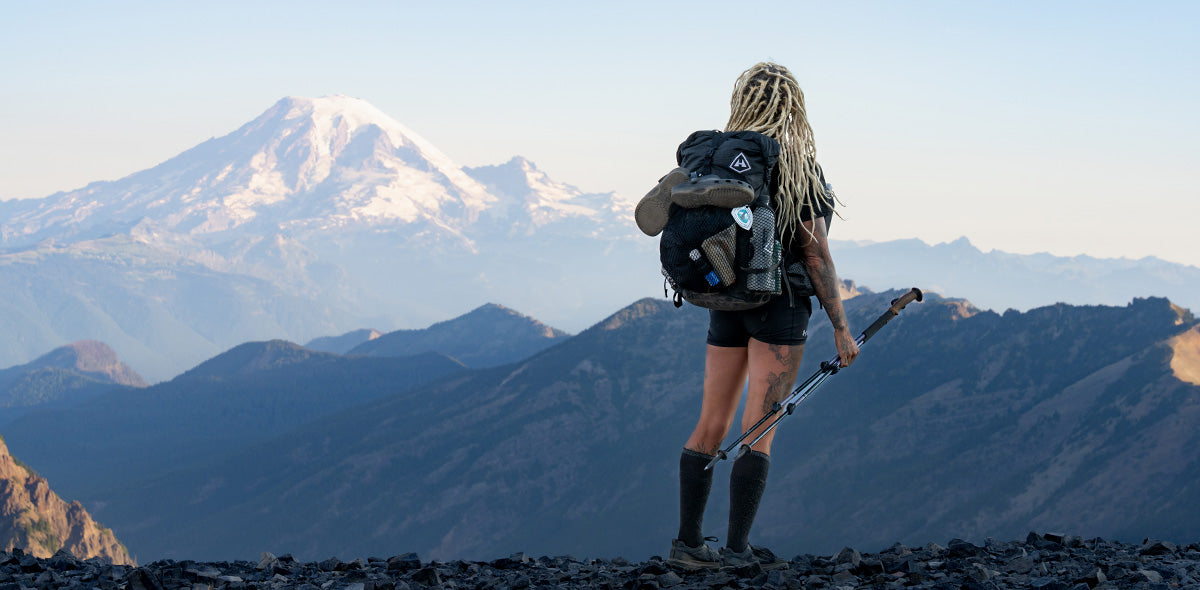 Person with a backpack and hiking poles standing on a mountain peak with snow-capped mountains in the background.
