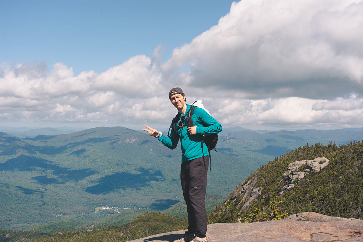Person standing on a mountain peak with a scenic view of clouds and mountains.