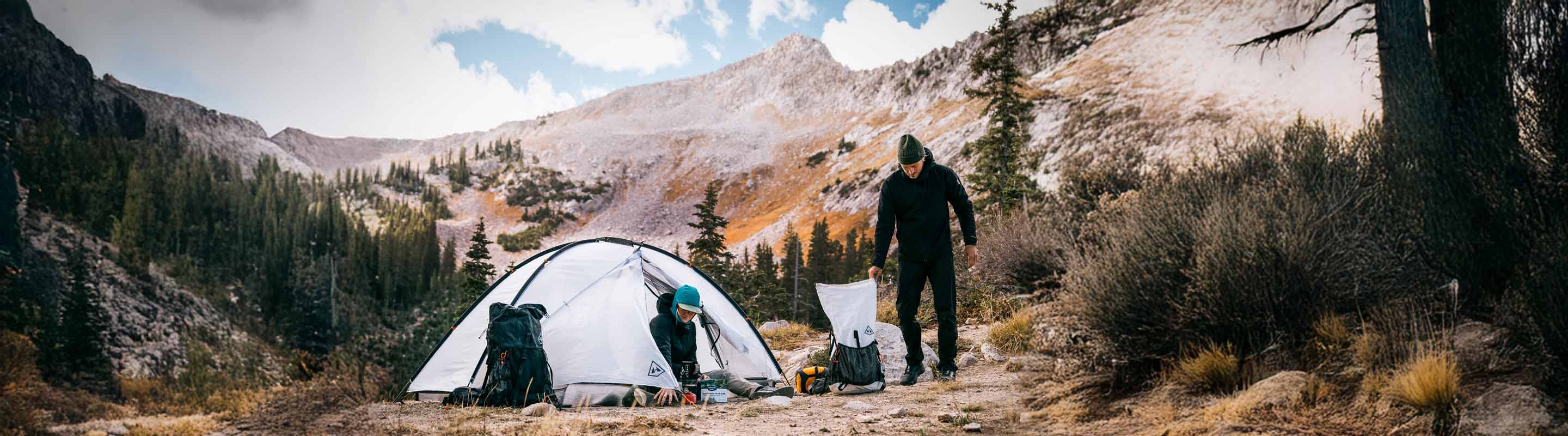 Two people in a camping area with a tent in a mountainous area with trees and rocks.