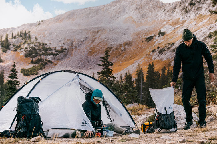 Two people in a camping area in a mountainous area with a tent, backpacks and gear.
