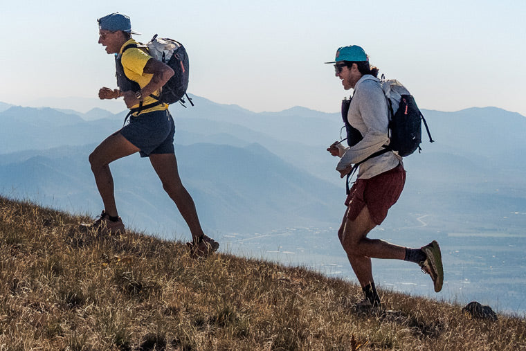Two people running on a grassy hill carrying backpacks with mountains in the background