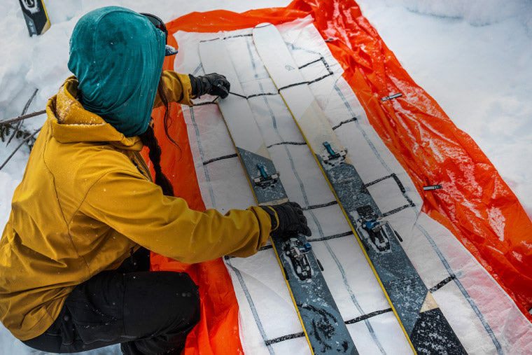Person preparing skis on an orange mat with snow in the background